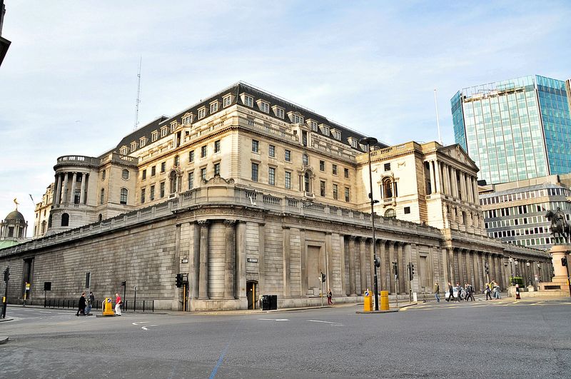 The Bank of England building on Threadneedle Street in London, a grand neoclassical stone facade with columns, seen from street level against a partly cloudy sky