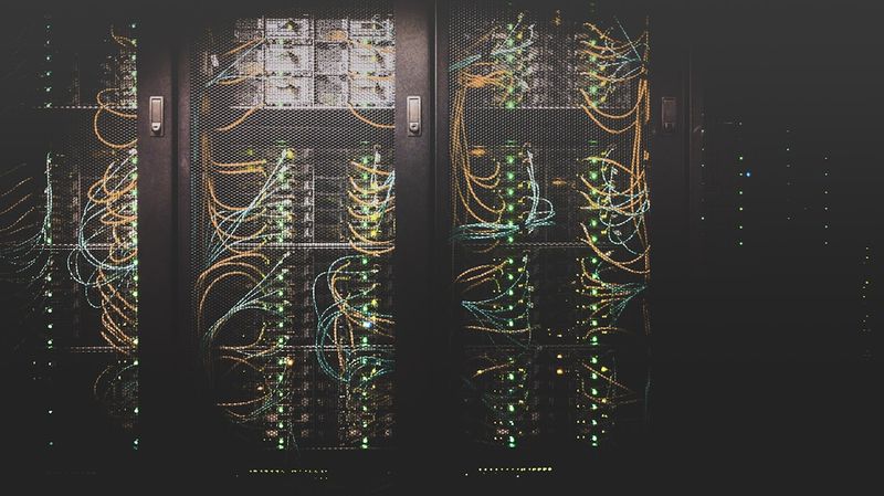 Dark server room with racks of networking equipment illuminated by green indicator lights and tangled cables