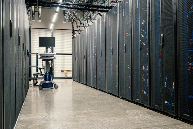 A long corridor of dark server racks inside a modern data center facility, with overhead cable management and a monitoring station on wheels