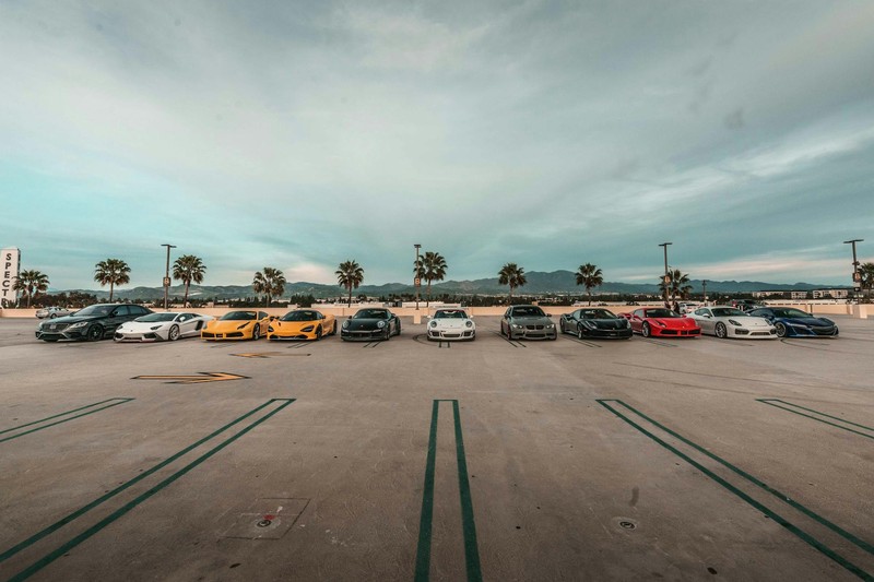 Lineup of luxury sports cars parked in a row on an open lot under a dramatic cloudy sky with palm trees in the background