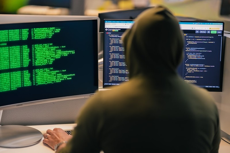 A hacker in a hoodie working at a desk with multiple computer screens displaying code and data